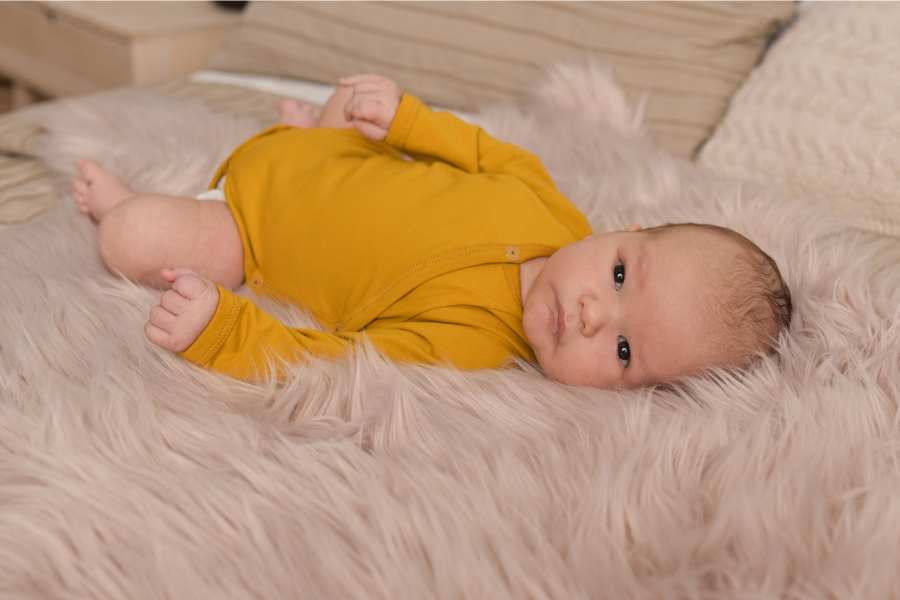 A small baby in a baby yellow bodysuit lies on artificial fur with high hair